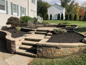 Stone steps and retaining walls surround tiered garden beds with fresh mulch and young plants next to a house. Orange markers are placed in the soil.