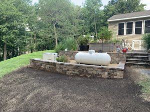 A backyard with a propane tank on a stone retaining wall, beside a house. The area features plants and a patch of bare soil, with trees in the background.