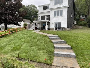 A white multi-story house with a freshly laid sod lawn. A stone pathway with steps leads to the front entry. A wooden fence borders the left side of the yard.