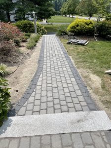 A paved stone walkway bordered by grass and plants, leading to a grassy area with trees in the background. A small stone structure is on the left.