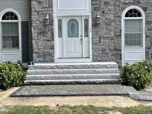 Front entrance with gray stone steps leading to a white door, flanked by two arched windows. Stone path in front. Bushes to the sides of the steps.