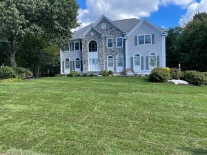 A large two-story house with stone and white siding, surrounded by a green lawn and trees, under a partly cloudy sky.