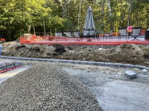 Construction site with a pile of gravel, an incomplete retaining wall, and a deck with patio furniture and an umbrella, surrounded by orange safety netting.