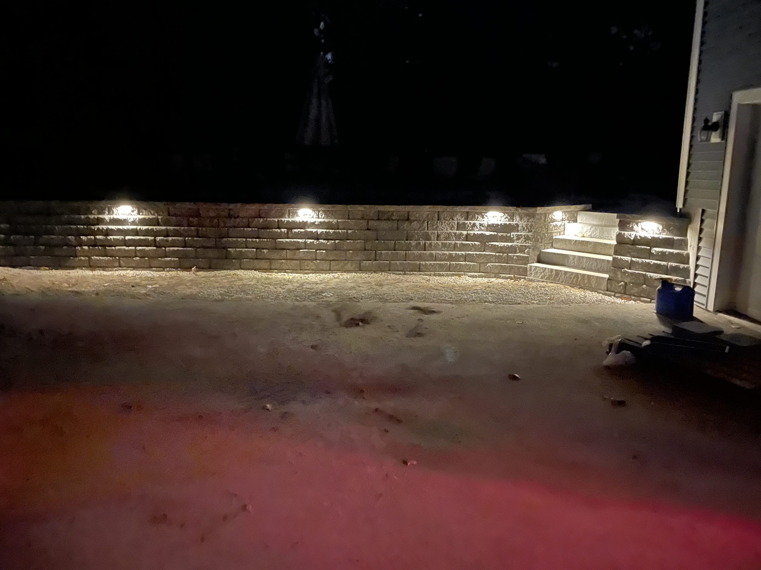 Dimly lit stone retaining wall with recessed lights, stairs on the right, adjacent to a building.