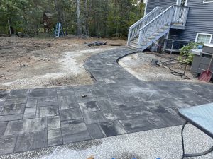 A newly laid stone pathway curves from a staircase to a patio area beside a house, with a ladder and trees in the background.