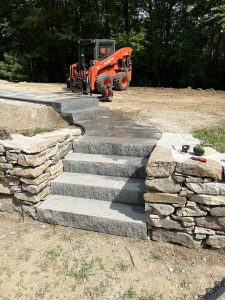 Stone stairs lead up to a partly cleared dirt area. A small orange skid steer loader is parked nearby, and some tools are on a ledge.