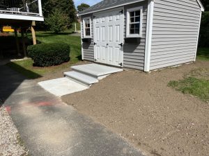 A gray shed with double doors sits on a concrete pad. Freshly laid dirt surrounds the structure. A small set of steps leads to the entry. Bushes and grass are visible in the background.