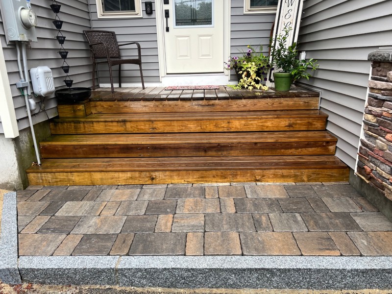 Wooden steps with flower pots lead to a white door, bordered by gray siding and decorative stonework. A paver patio lies in the foreground.