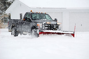 A snowplow truck is clearing a snowy driveway in front of a garage during a snowstorm.