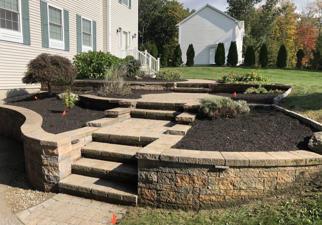 Stone steps and retaining walls surround tiered garden beds with fresh mulch and young plants next to a house. Orange markers are placed in the soil.