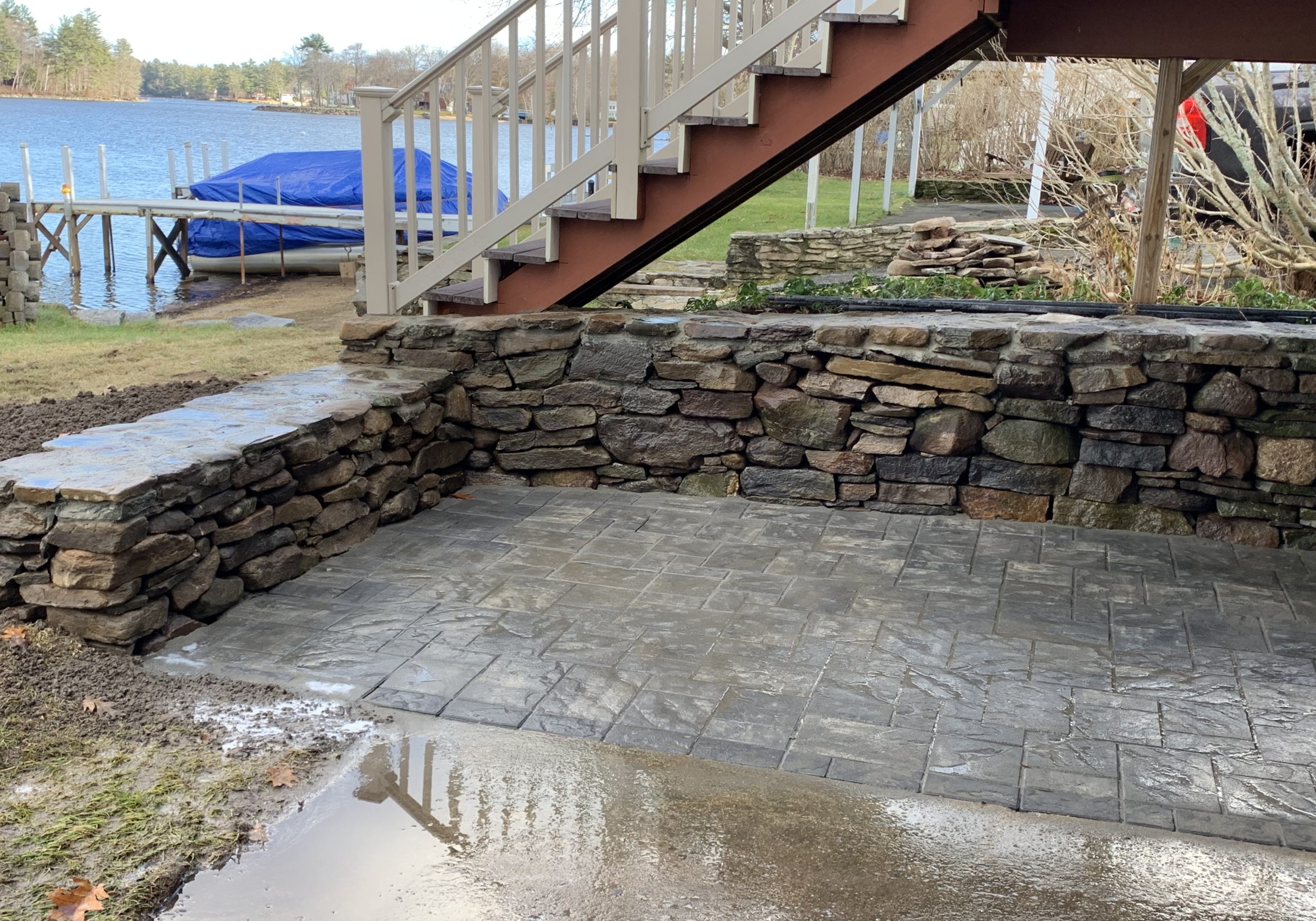 Stone patio under stairs near a lake with a retaining wall. The ground is wet, and there's a covered boat dock in the background.