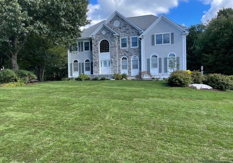 A large two-story house with stone and white siding, surrounded by a green lawn and trees, under a partly cloudy sky.